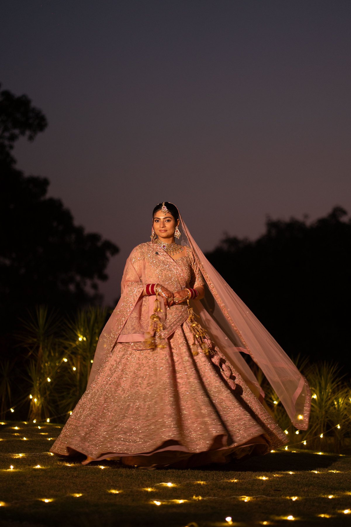 A bride in a flowing blush lehenga beneath a dusky sky