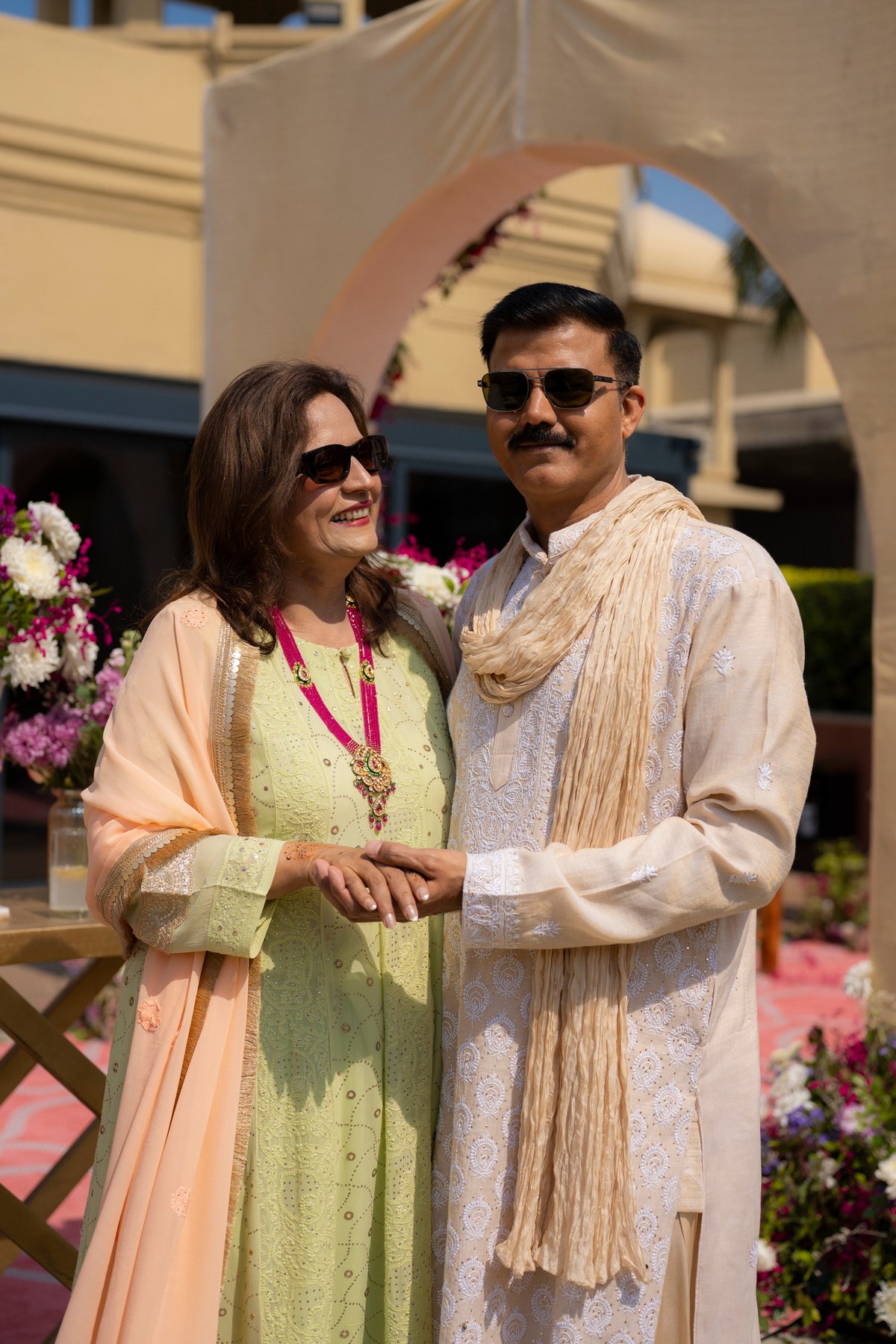 A smiling pair in pastel celebration attire beneath a flower-filled arch